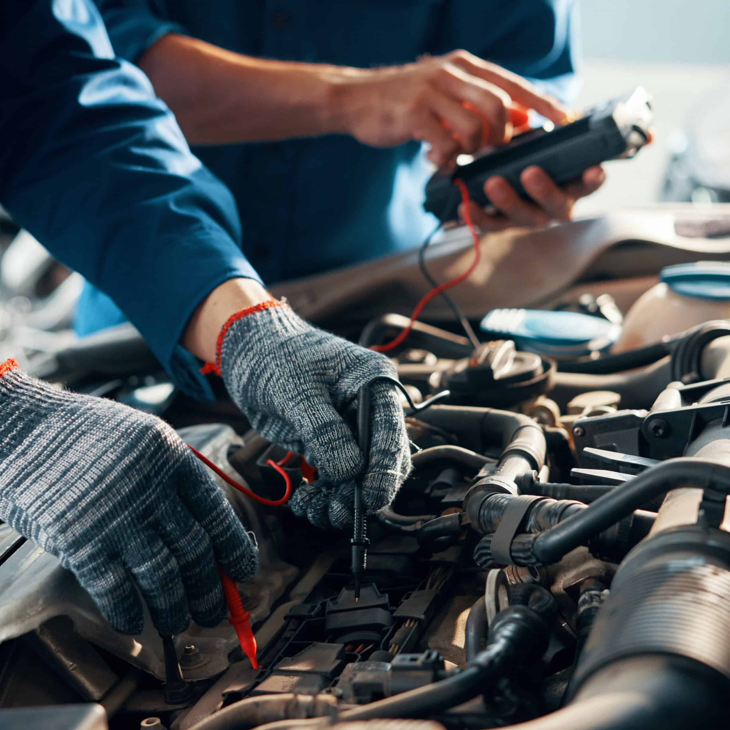 Deux mécaniciens testent un moteur de voiture avec un multimètre. L'un porte des gants et utilise la sonde, l'autre tient l'appareil pour le diagnostic électrique.