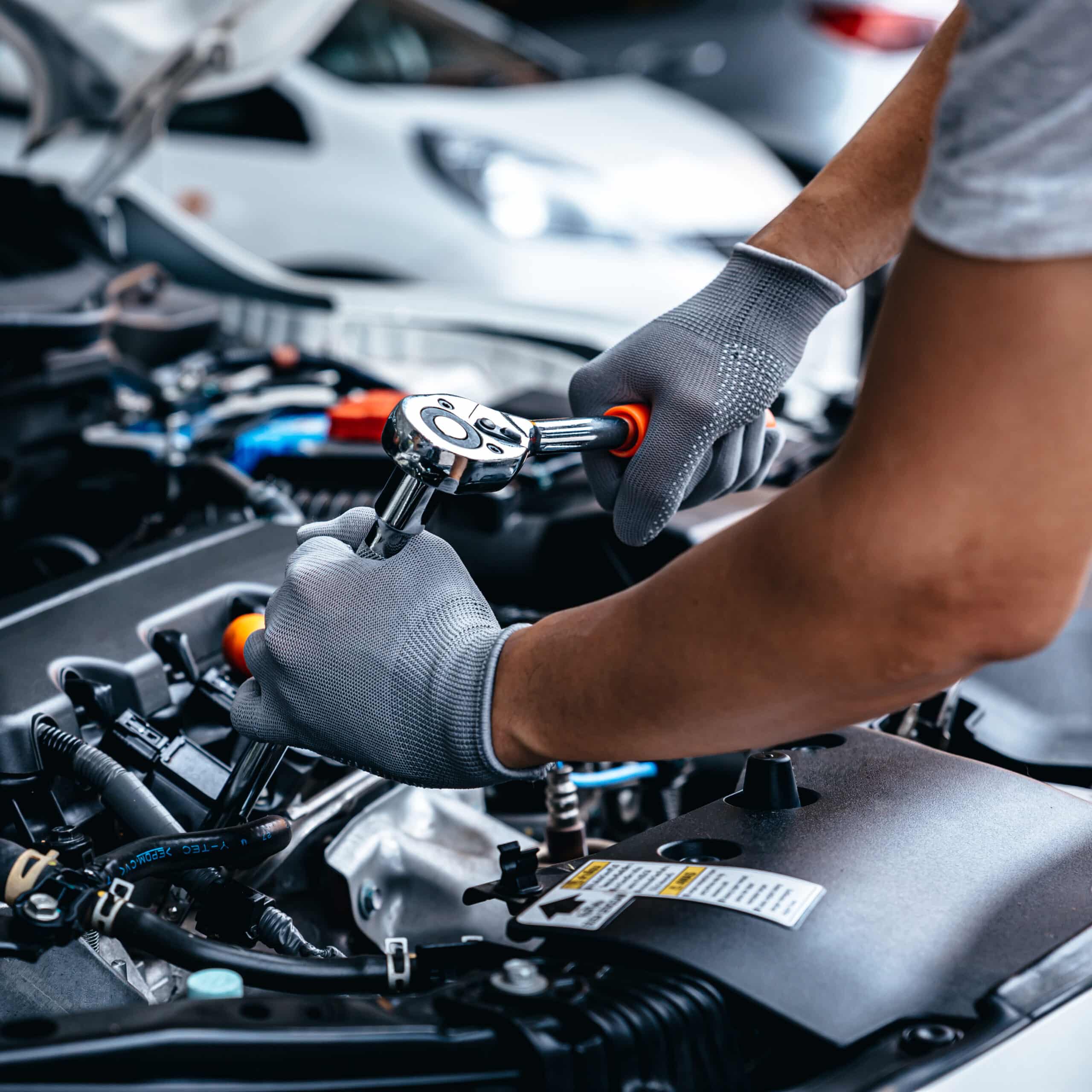 Mécanicien ganté répare un moteur de voiture avec une clé à cliquet. Détail 'AWARNING' et tuyau 'Y-TEC' visibles sur le moteur.