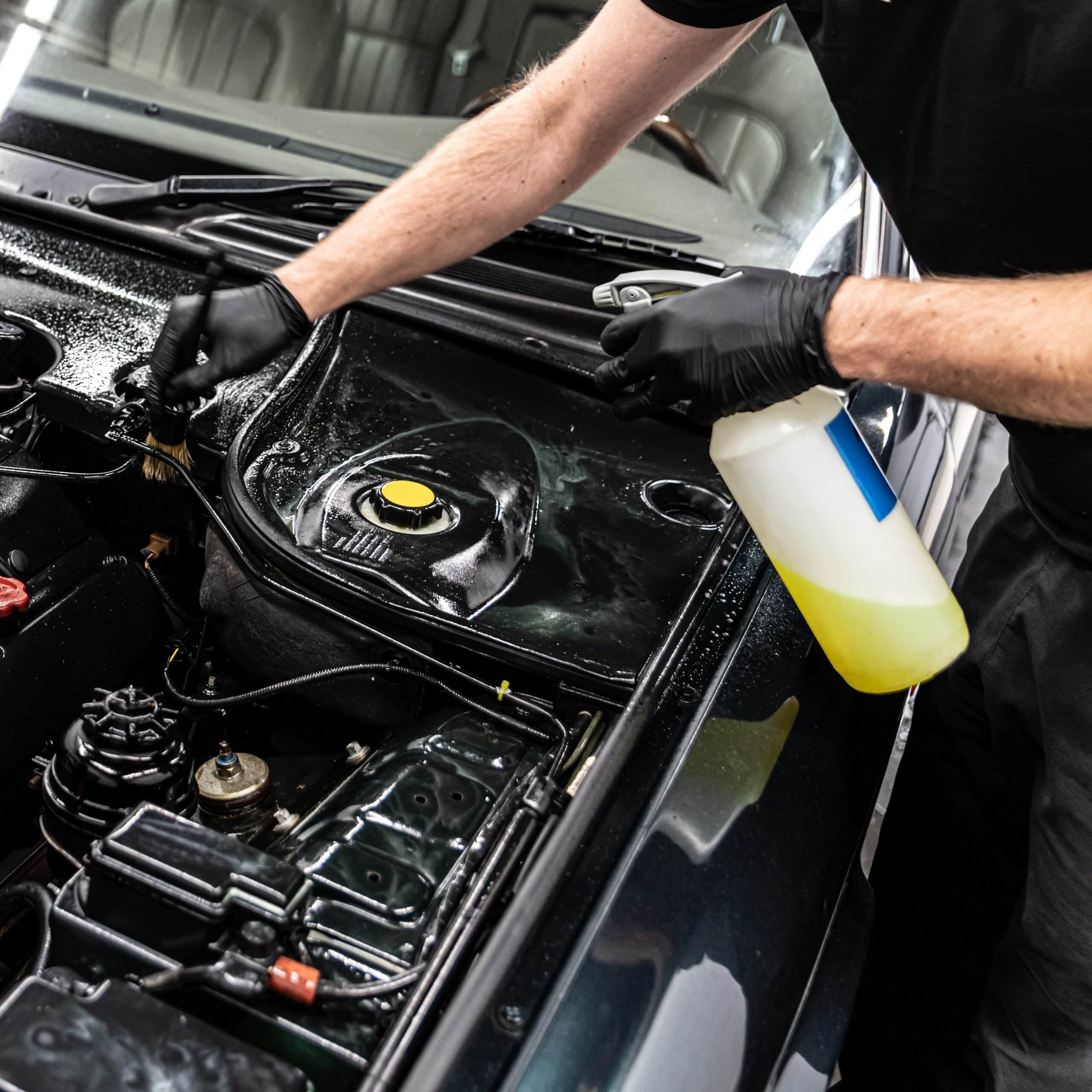 Nettoyage professionnel d'un compartiment moteur de voiture. Mains gantées appliquant un produit jaune avec brosse et pulvérisateur.