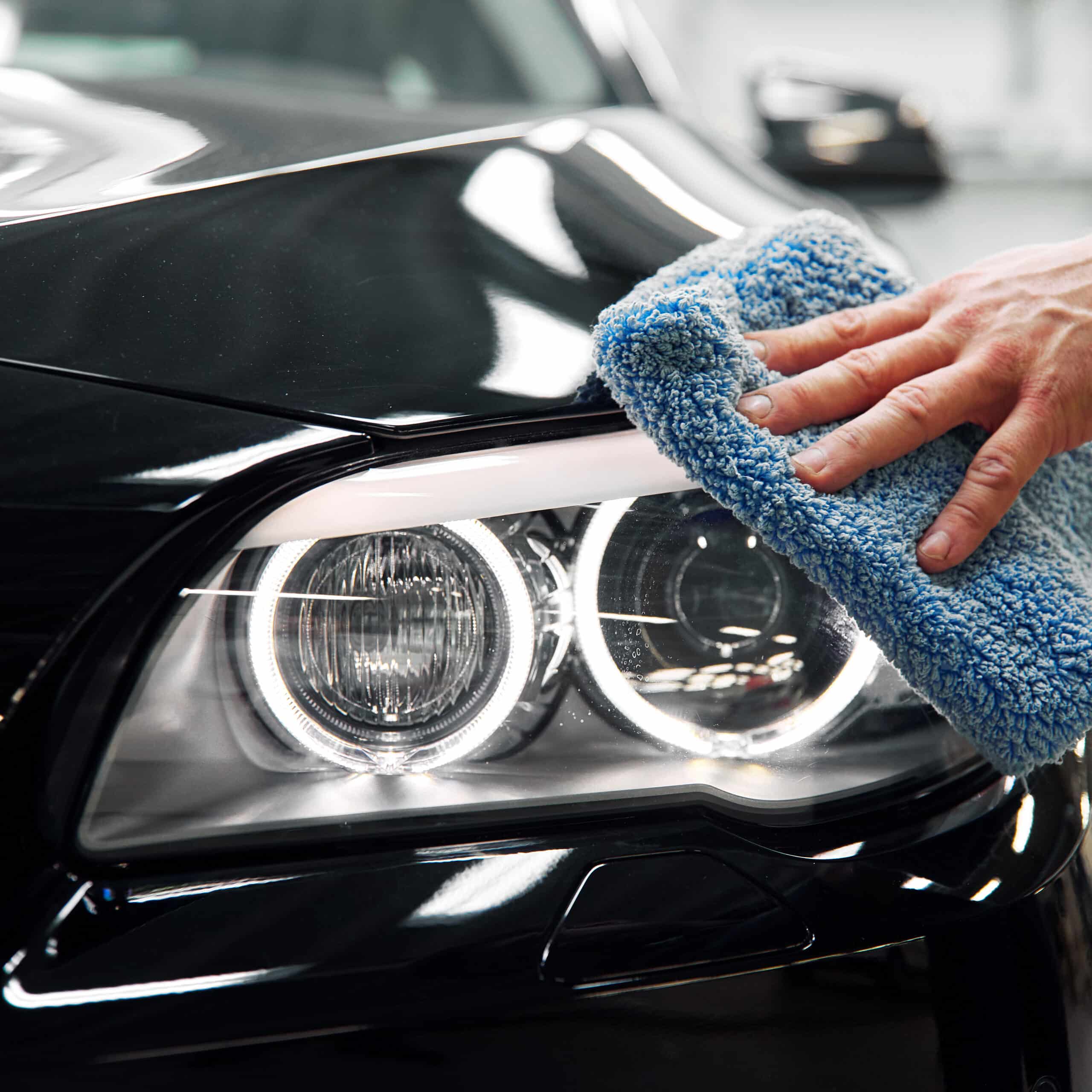 Main nettoyant le phare lumineux et la carrosserie noire brillante d'une voiture avec une serviette microfibre bleue douce.