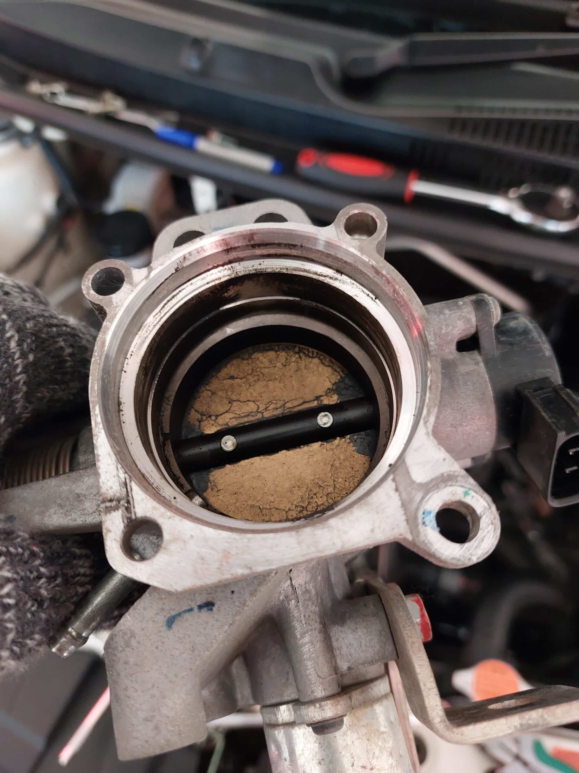 Papillon des gaz encrassé avec d'épais dépôts brunâtres craquelés sur le clapet, tenu par une main gantée. Indique un besoin urgent de nettoyage moteur.