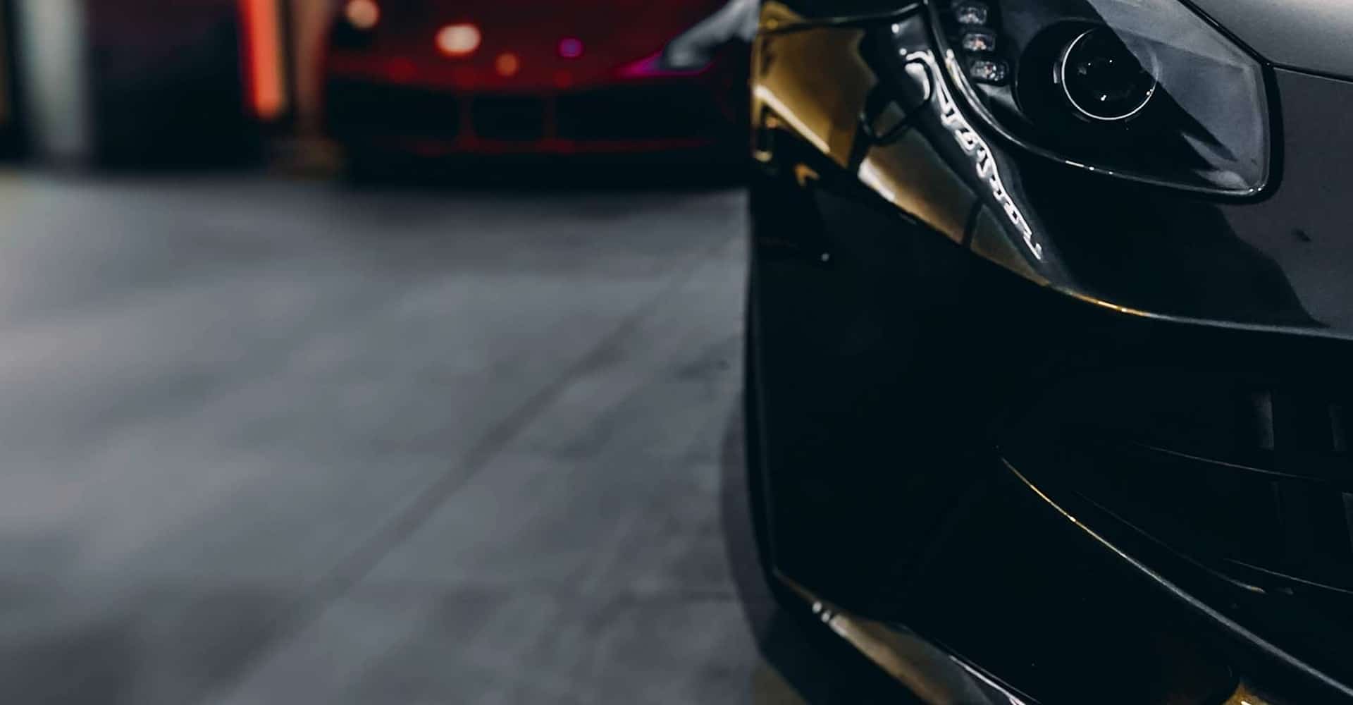 Avant d'une voiture de sport noire brillante, son phare LED détaillé en gros plan, dans un garage avec un véhicule rouge flou en arrière-plan.