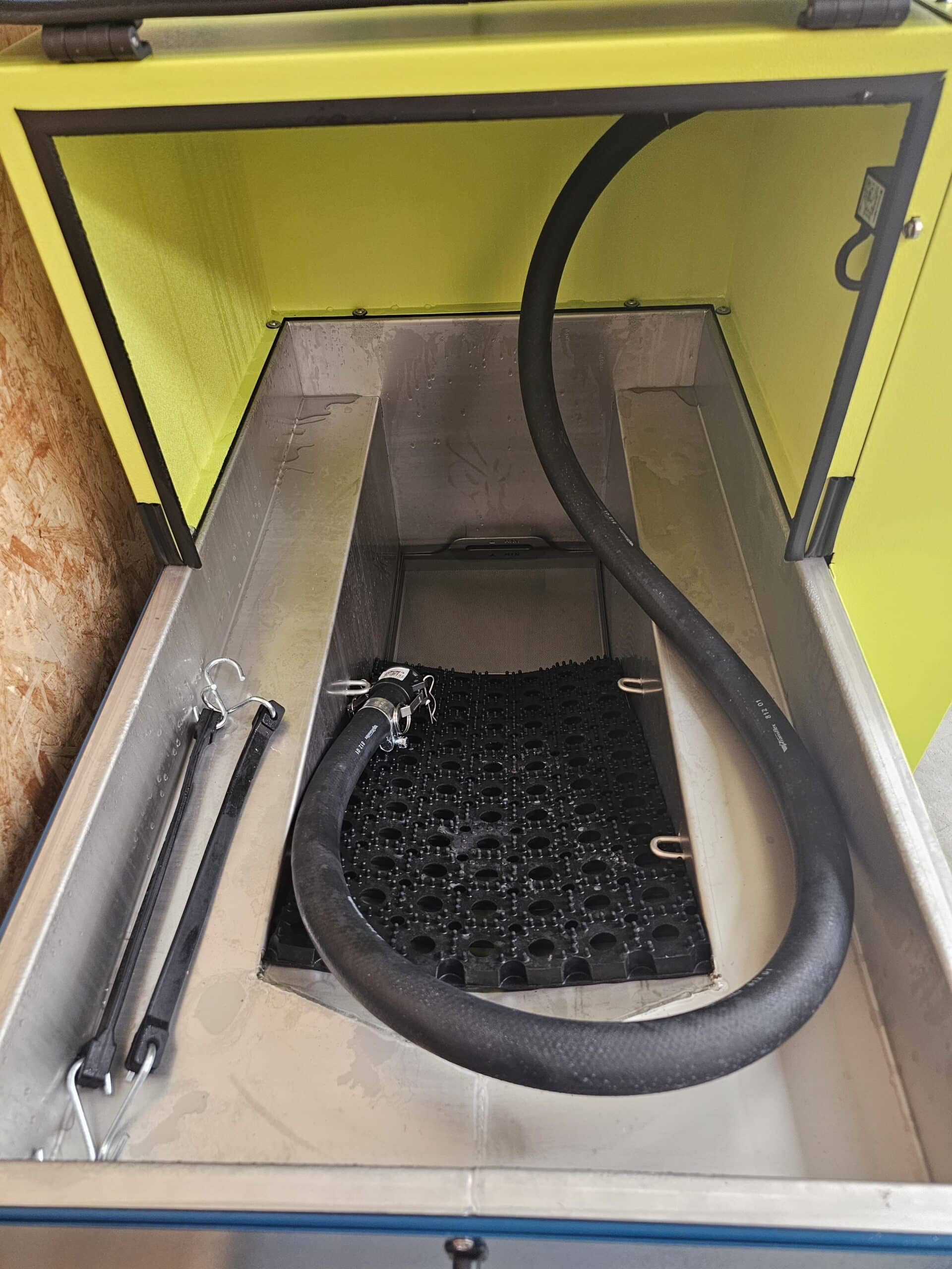 Intérieur d'une machine de nettoyage industrielle avec cuve inox, tuyau noir et tapis de protection en caoutchouc.